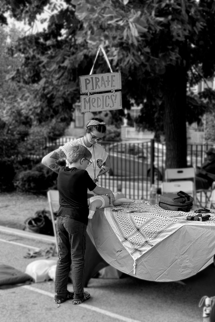 A black and white photo showing one of the pirate medics team members standing over a child as the child practices performing infant CPR on a manikin